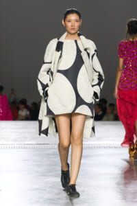 Model walking the runway in a white and black abstract-patterned coat over a matching dress, hands in pockets, focused expression.