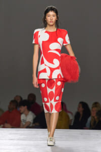 Model on a runway wearing a red and white abstract-print dress with matching skirt and a red fluffy bag over the arm