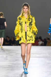 Model on a fashion runway wearing a bright yellow embroidered coat with black floral patterns and blue sneakers on the catwalk.
