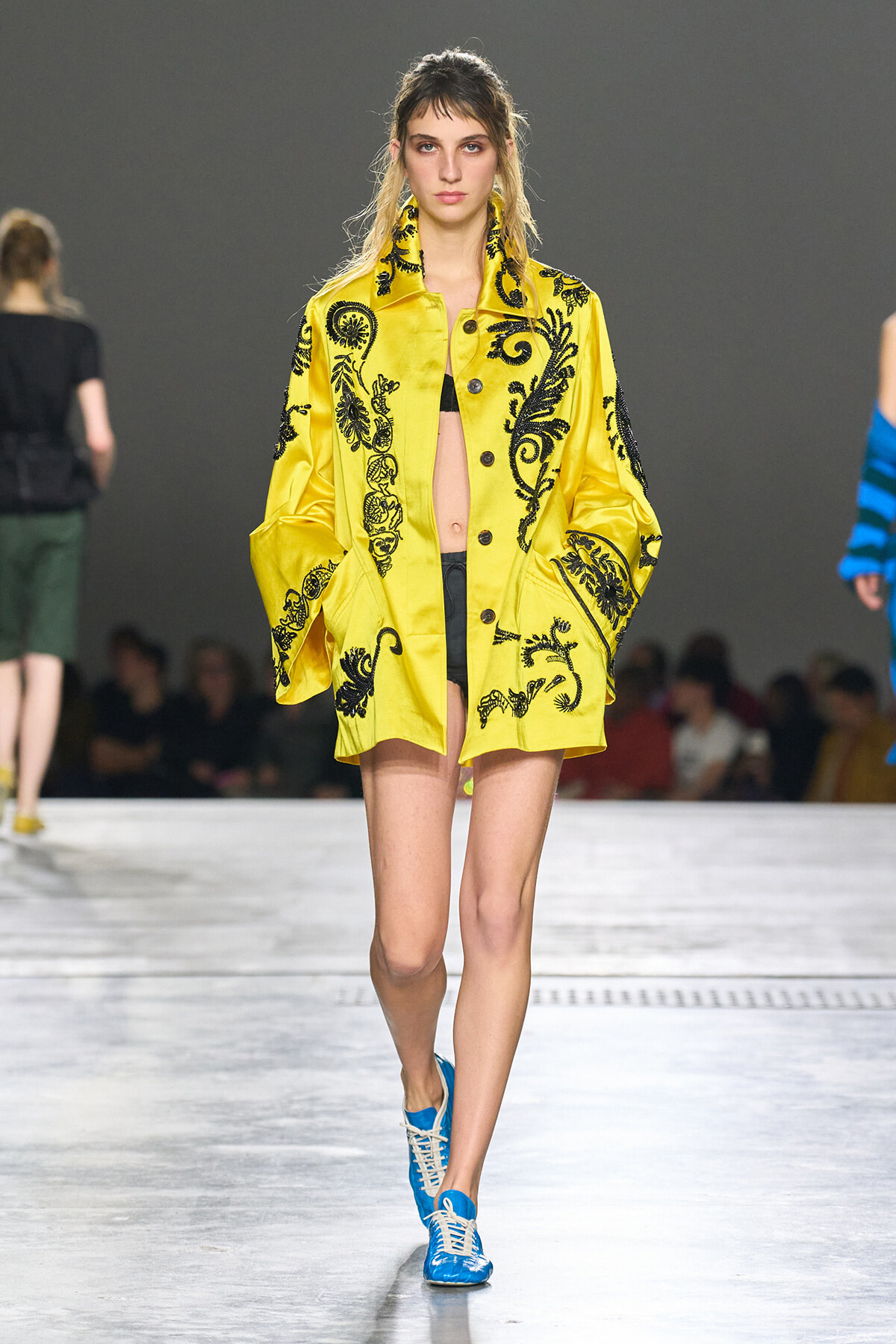 Model on a fashion runway wearing a bright yellow embroidered coat with black floral patterns and blue sneakers on the catwalk.