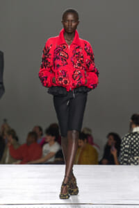 Model walking a runway in a bright pink embroidered bomber jacket with black floral embellishments and a black layered hem, paired with knee-length black shorts and patterned heels.