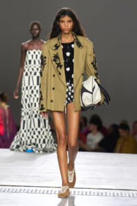 Runway model wearing a tan embroidered blazer over a black dress with white patterns, carrying a large white and black geometric tote bag.