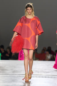 Model walking the runway in a short pink-orange geometric dress with oversized sleeves, carrying a bright red fluffy handbag and wearing gold strappy heels.
