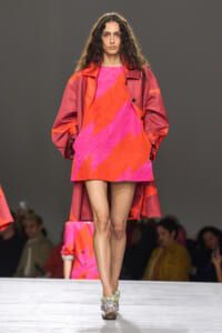 Model walks the runway in a pink and orange zigzag mini dress with a red oversized coat and jeweled heels, audience in the background