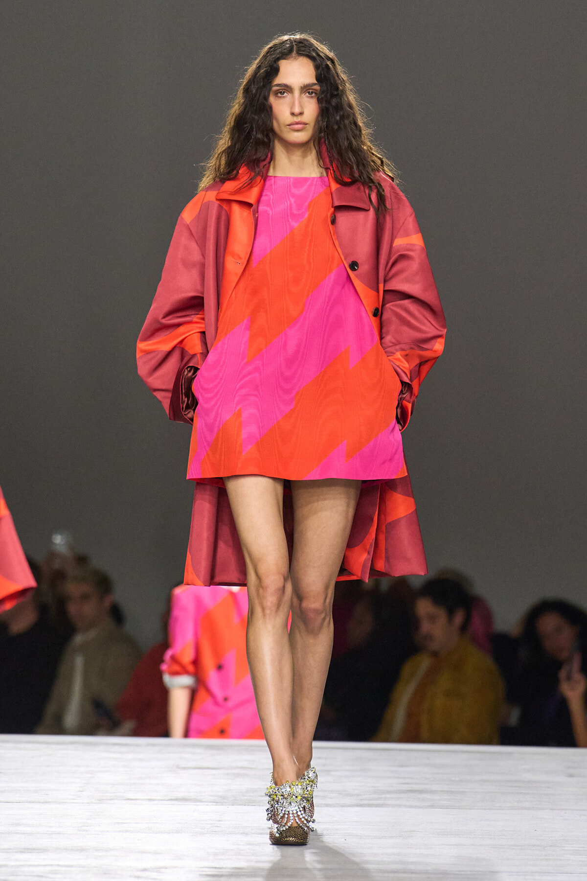Model walks the runway in a pink and orange zigzag mini dress with a red oversized coat and jeweled heels, audience in the background