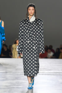 Model walking the runway in a dark gray polka-dot double-breasted coat with oversized lapels and bright blue sneakers.