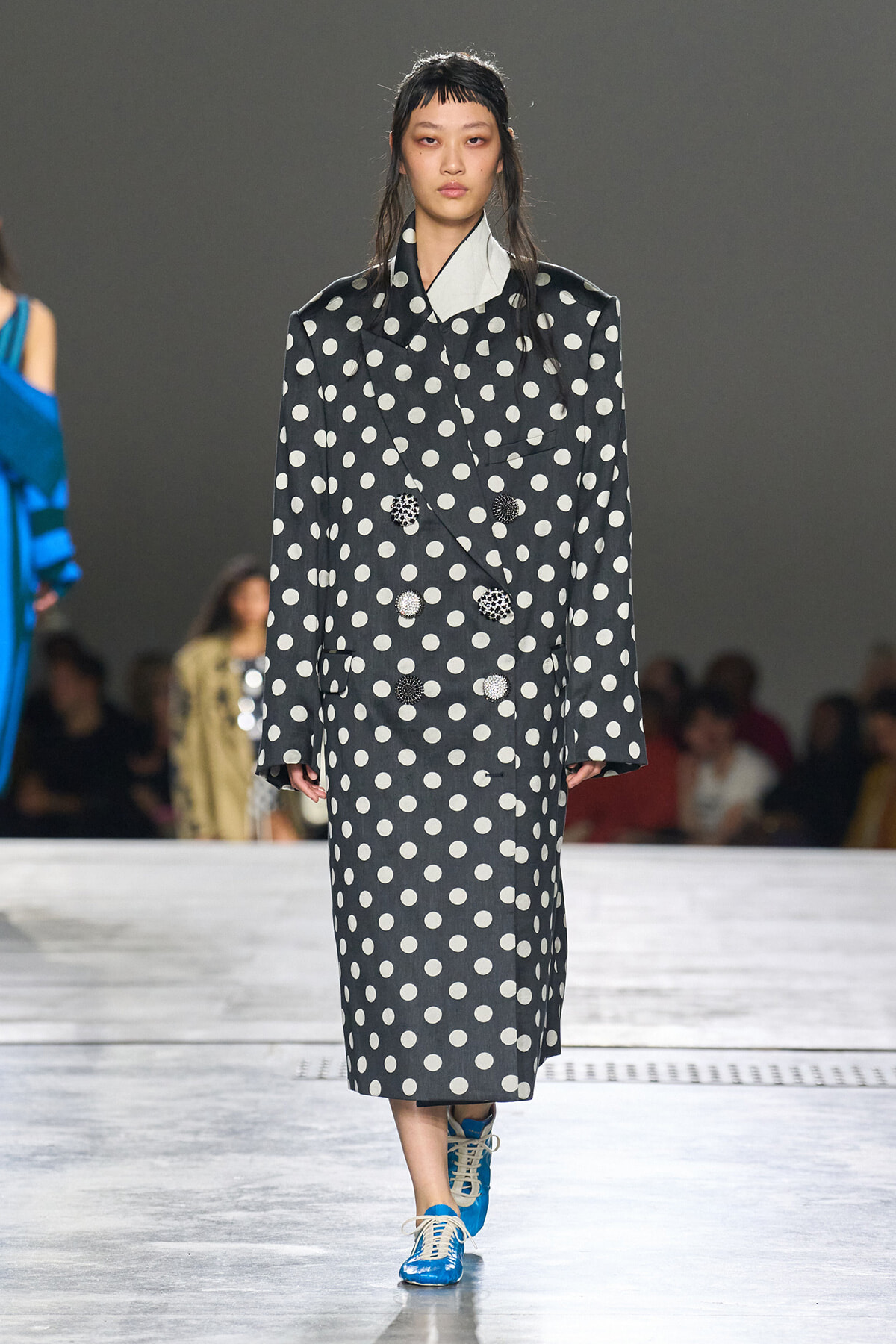 Model walking the runway in a dark gray polka-dot double-breasted coat with oversized lapels and bright blue sneakers.