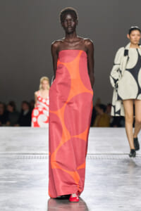 Model walks the runway in a strapless pink maxi dress with bold orange abstract shapes, paired with red shoes.