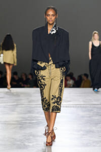 Model walks the runway in an oversized black blazer paired with beige cropped pants featuring black floral embroidery and brown tie-up heels.