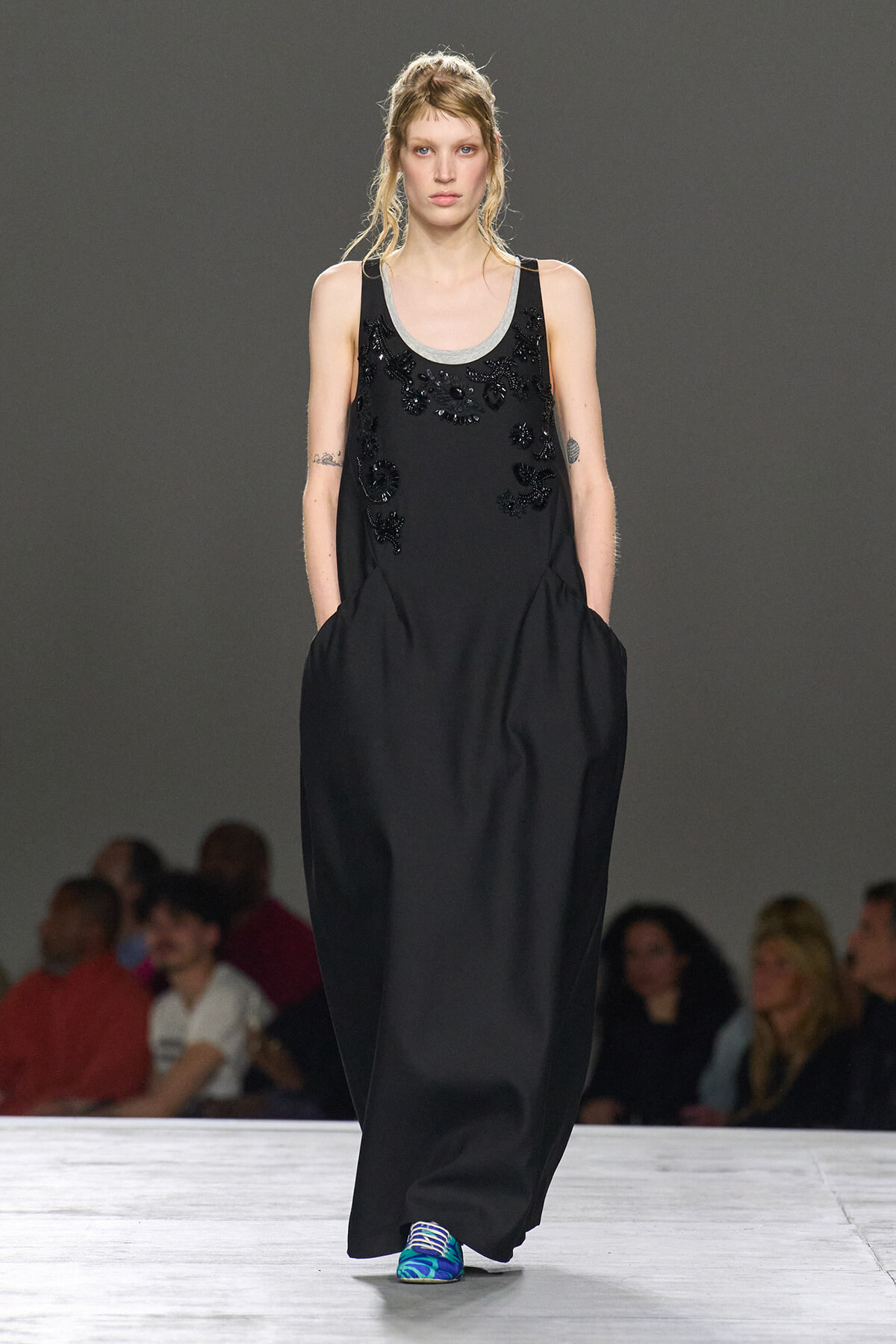 Model on a runway wearing a long black sleeveless dress with beaded floral appliqué and a gray neckline, pockets at the sides, paired with bright sneakers.