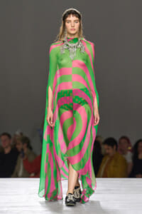 Model walking a runway in a sheer neon green and pink abstract-print gown with a large crystal necklace.