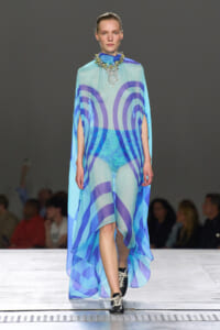 Model walks a runway wearing a sheer cyan cape dress with blue circular patterns and matching shorts, accessorized by a large jeweled necklace.