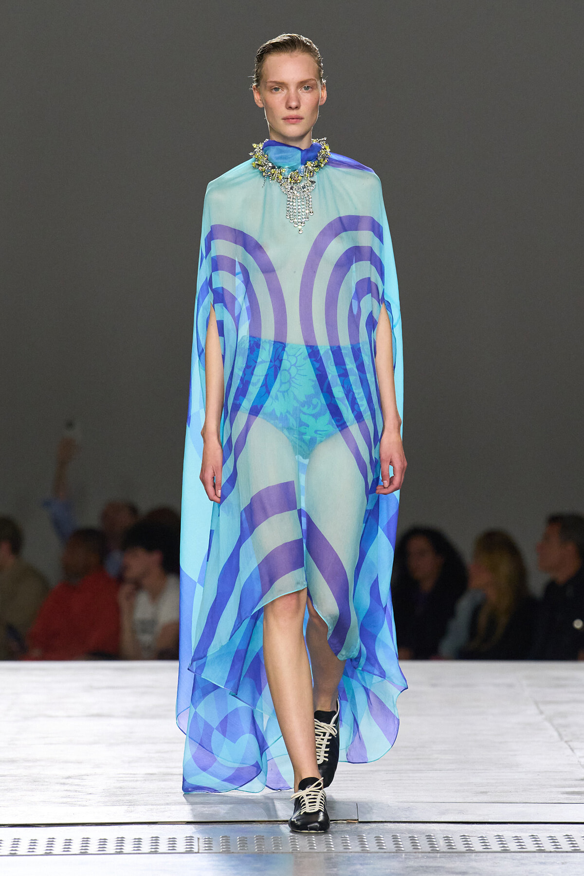 Model walks a runway wearing a sheer cyan cape dress with blue circular patterns and matching shorts, accessorized by a large jeweled necklace.