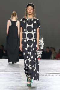 Runway model wearing a black-and-white polka-dot maxi dress with a statement silver necklace and green fringe heels.