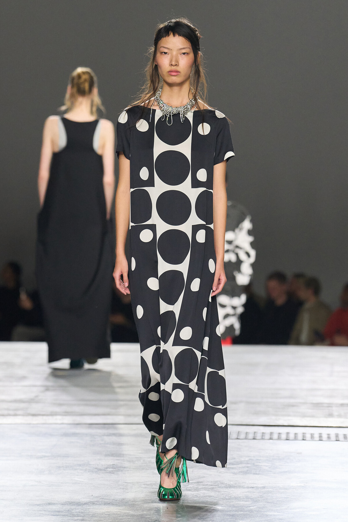 Runway model wearing a black-and-white polka-dot maxi dress with a statement silver necklace and green fringe heels.
