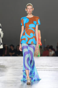 Model walking a runway in a bold color-block dress with brown, blue, and turquoise abstract patterns.
