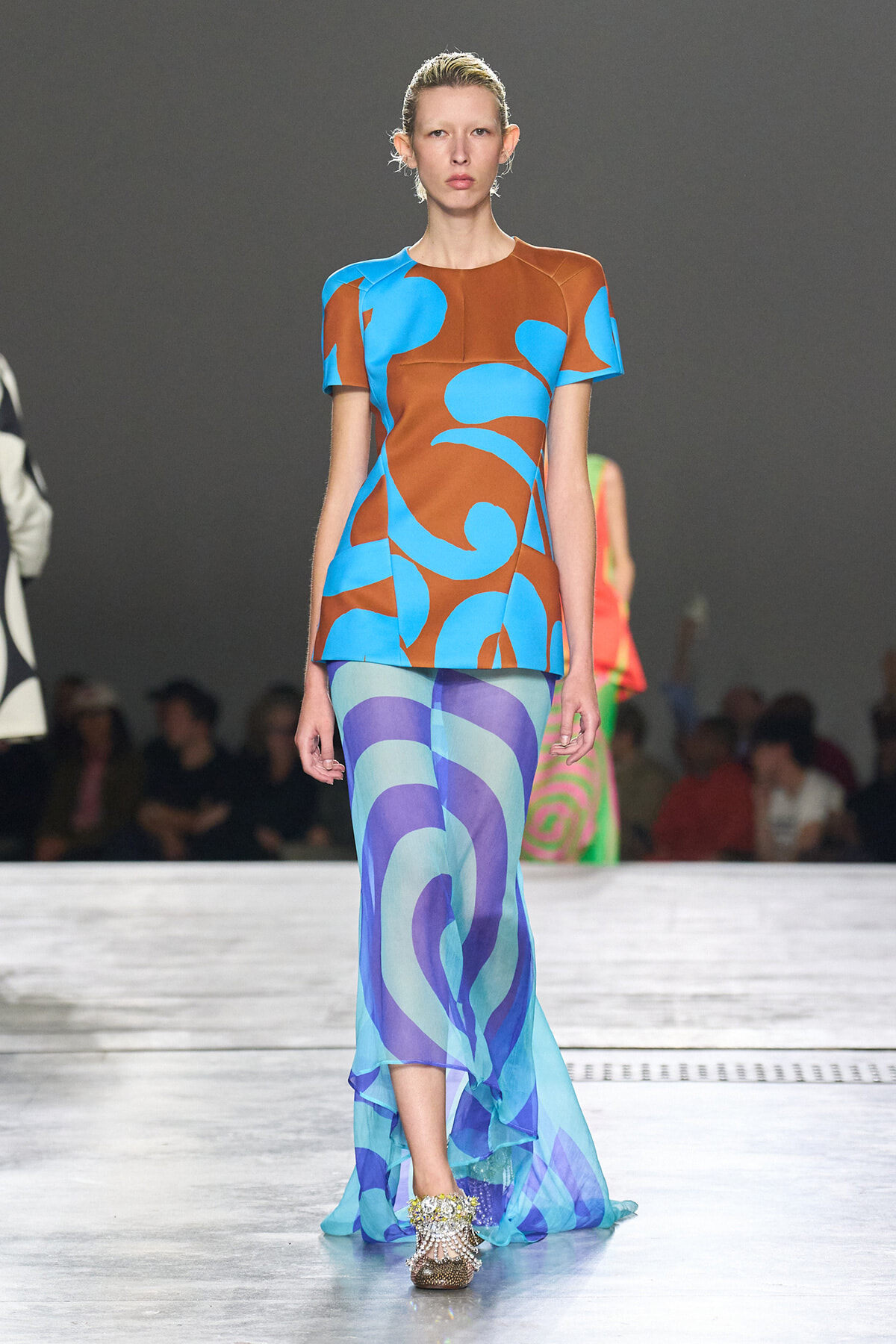 Model walking a runway in a bold color-block dress with brown, blue, and turquoise abstract patterns.