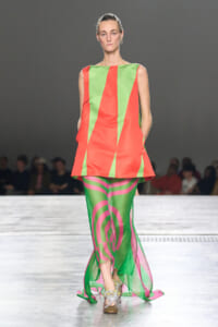 Model on a runway wearing a neon coral dress with lime-green geometric panels and matching swirling green and pink pants, plus glittery shoes.