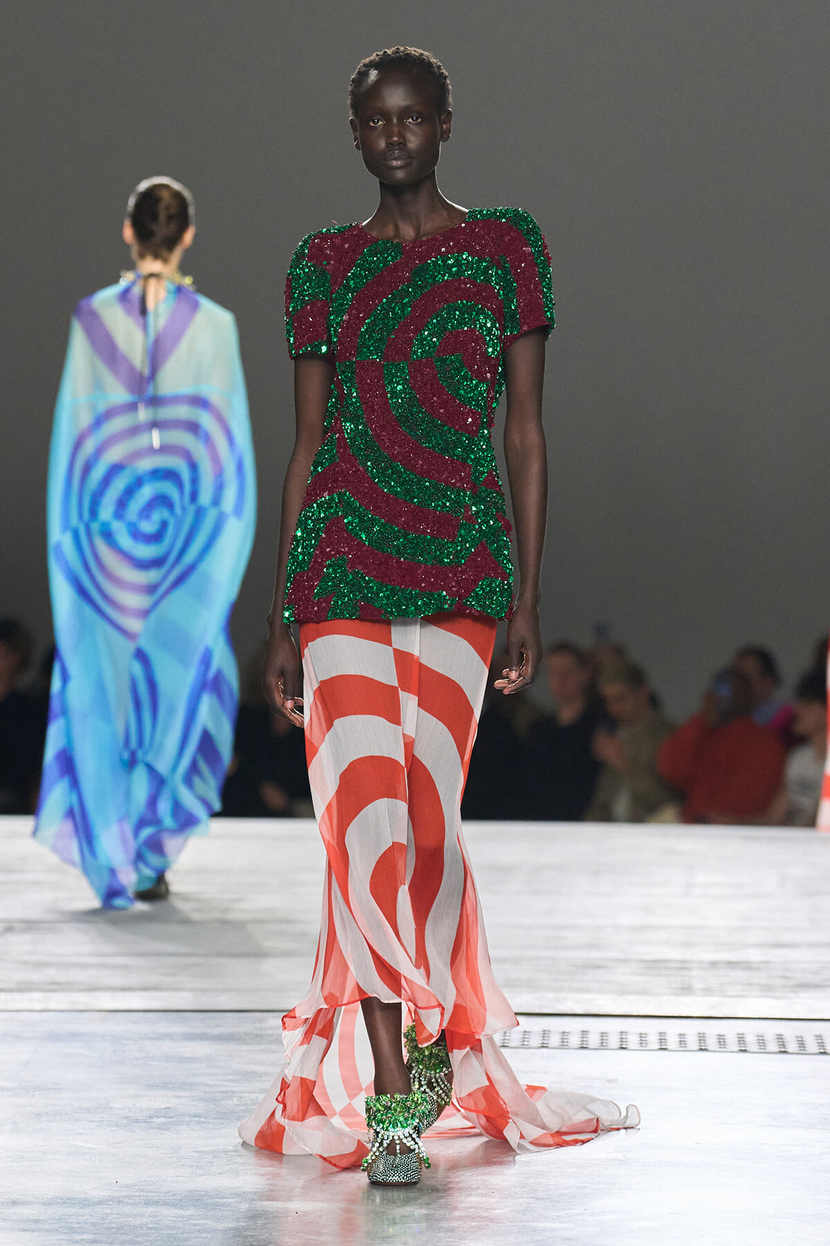 Model walking a runway in a green-and-burgundy sequined top with an orange-and-white swirl skirt, paired with green embellished shoes; audience in background.