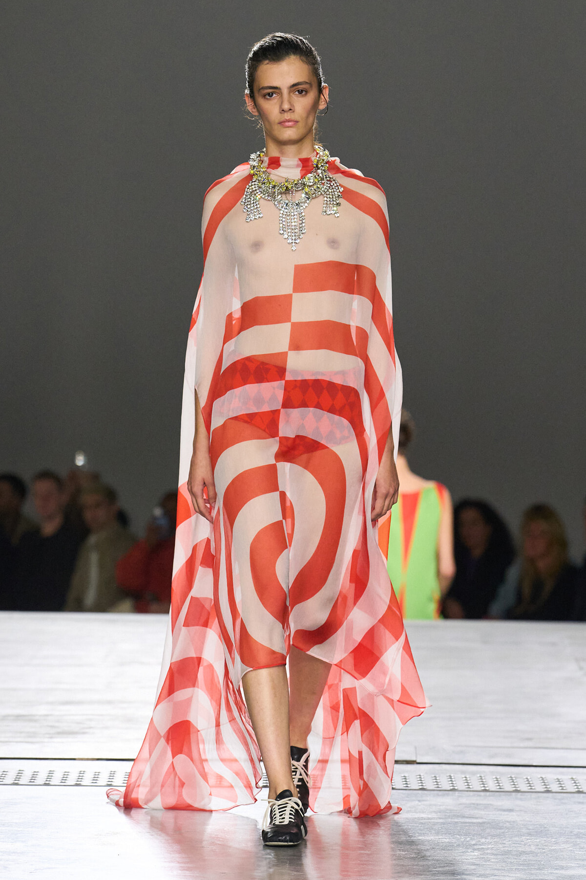 Model walks the runway wearing a sheer red-and-white geometric cape dress with a bold statement jeweled necklace; audience blurred in the background.