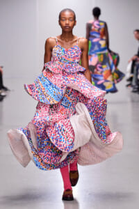 Model walking a runway in a bright, multicolored abstract-print gown with oversized, swirling layers; pink socks and brown shoes.
