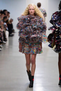 Model walking a runway in a voluminous multicolored ruffled dress with black ankle boots.