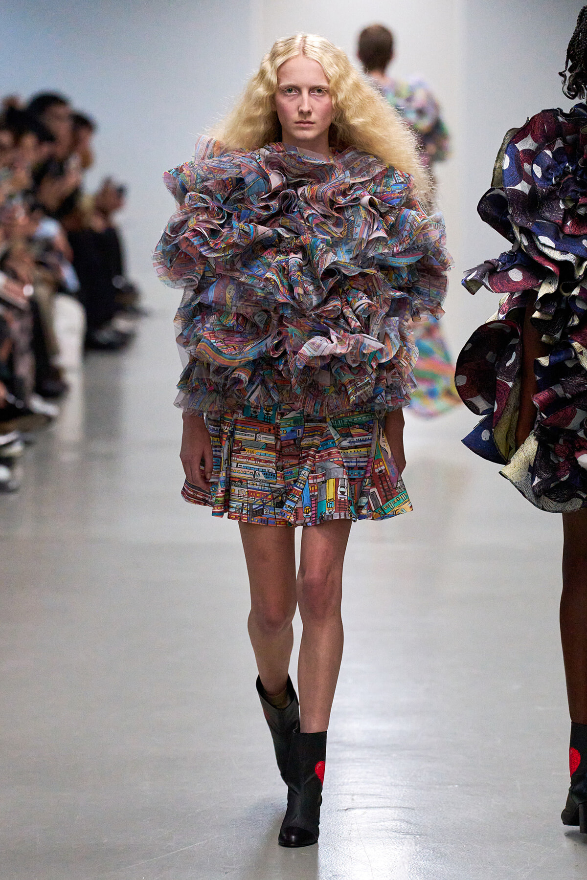 Model walking a runway in a voluminous multicolored ruffled dress with black ankle boots.