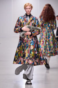 Model on a runway wearing a bright, abstract patterned coat while holding a gray fur stole; blurred backstage figure in background.