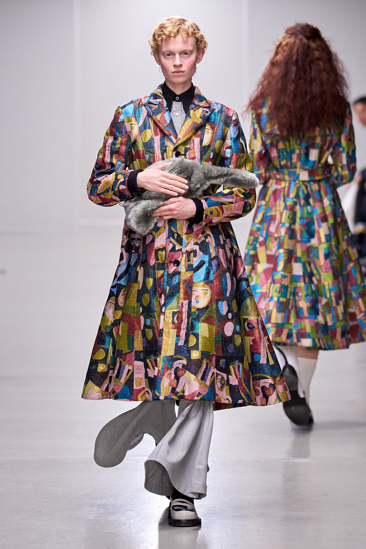 Model on a runway wearing a bright, abstract patterned coat while holding a gray fur stole; blurred backstage figure in background.
