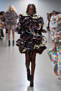 Model walks the runway in a voluminous, multi-layered ruffle dress with dark boots and braided hair.