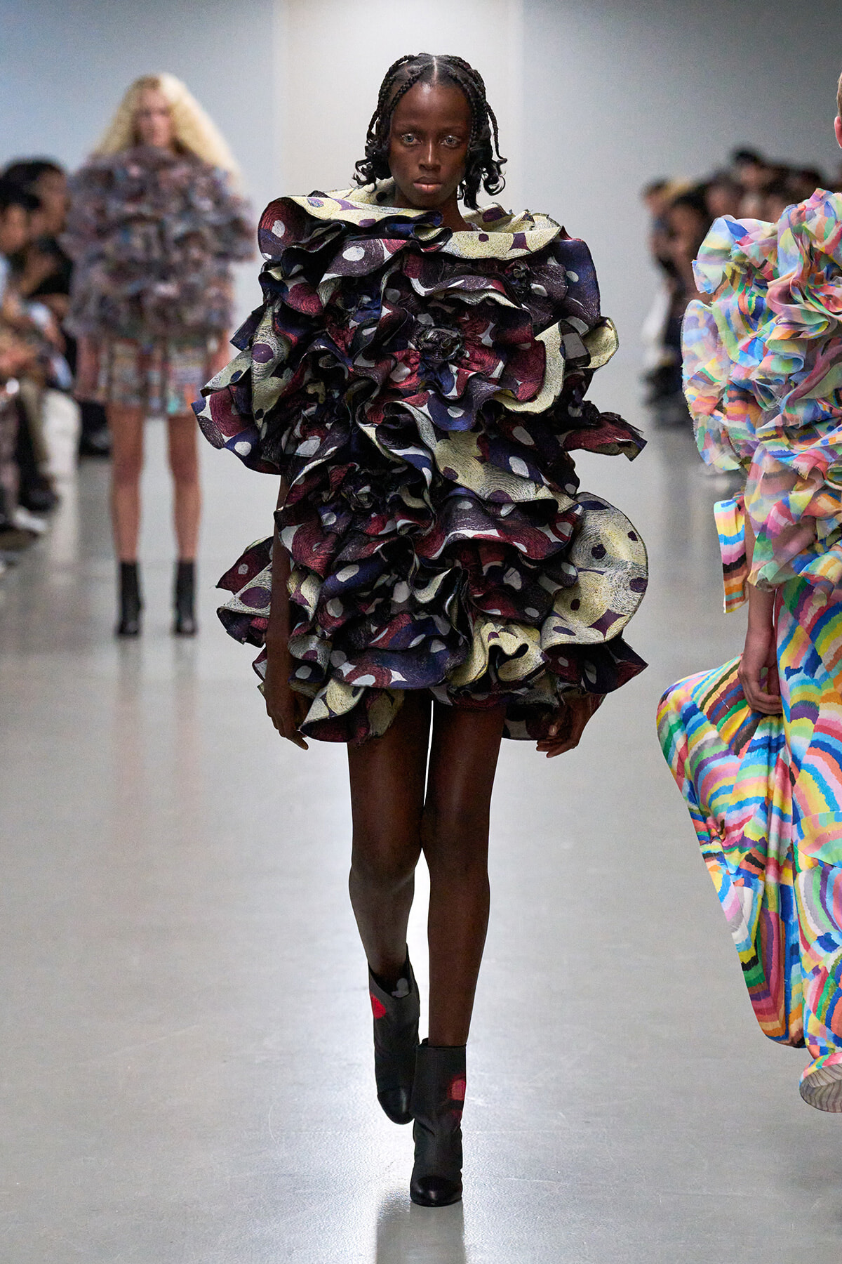 Model walks the runway in a voluminous, multi-layered ruffle dress with dark boots and braided hair.