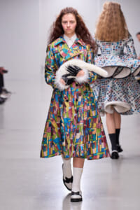 Model walks the runway in a vibrant abstract-patterned coat, black gloves with white fur trim, and a white fluffy accessory in her arms.