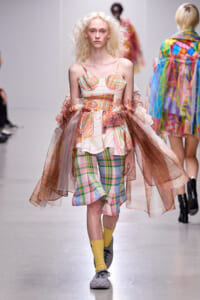 Runway model wearing a multicolor plaid dress with a sheer, voluminous cape and yellow socks during a fashion show