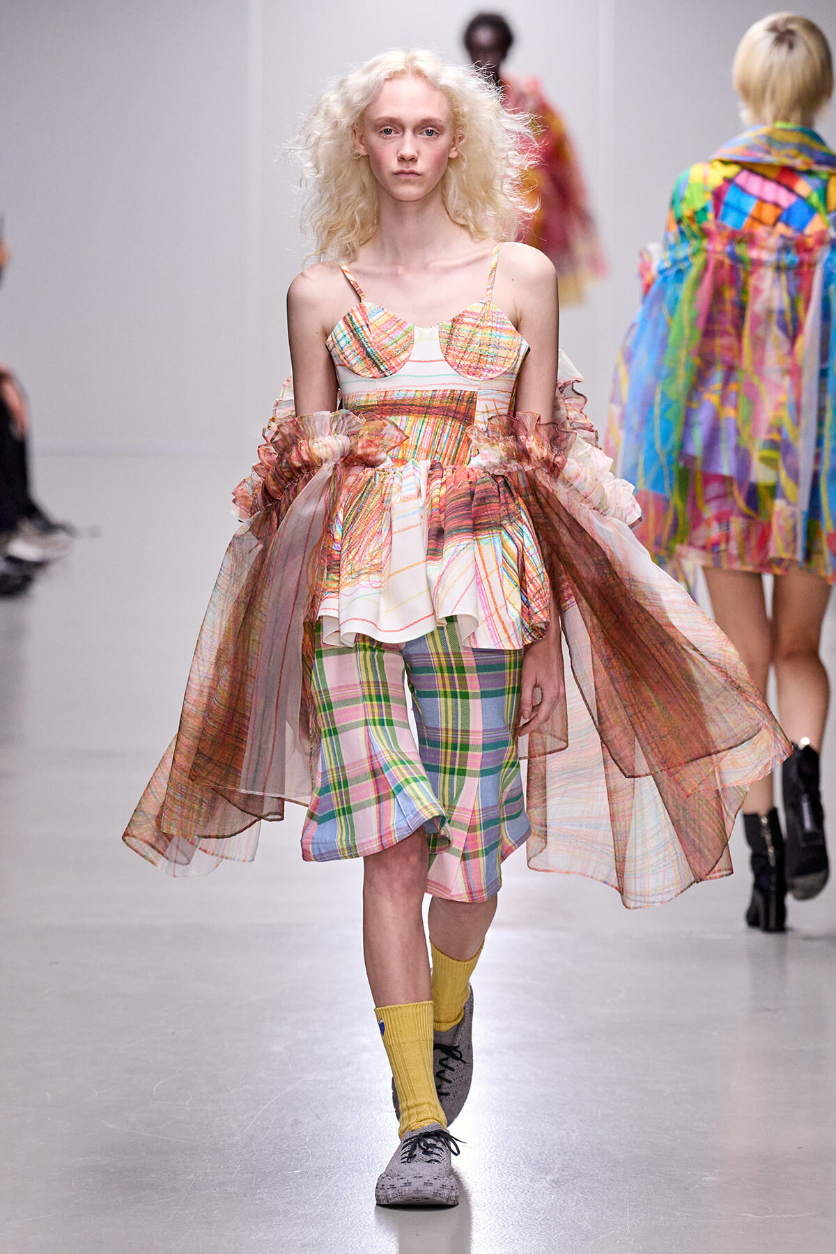 Runway model wearing a multicolor plaid dress with a sheer, voluminous cape and yellow socks during a fashion show