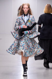 Model on a runway wearing a multicolored mosaic-pattern suit and dress, holding a black fluffy scarf with a badge on the chest.