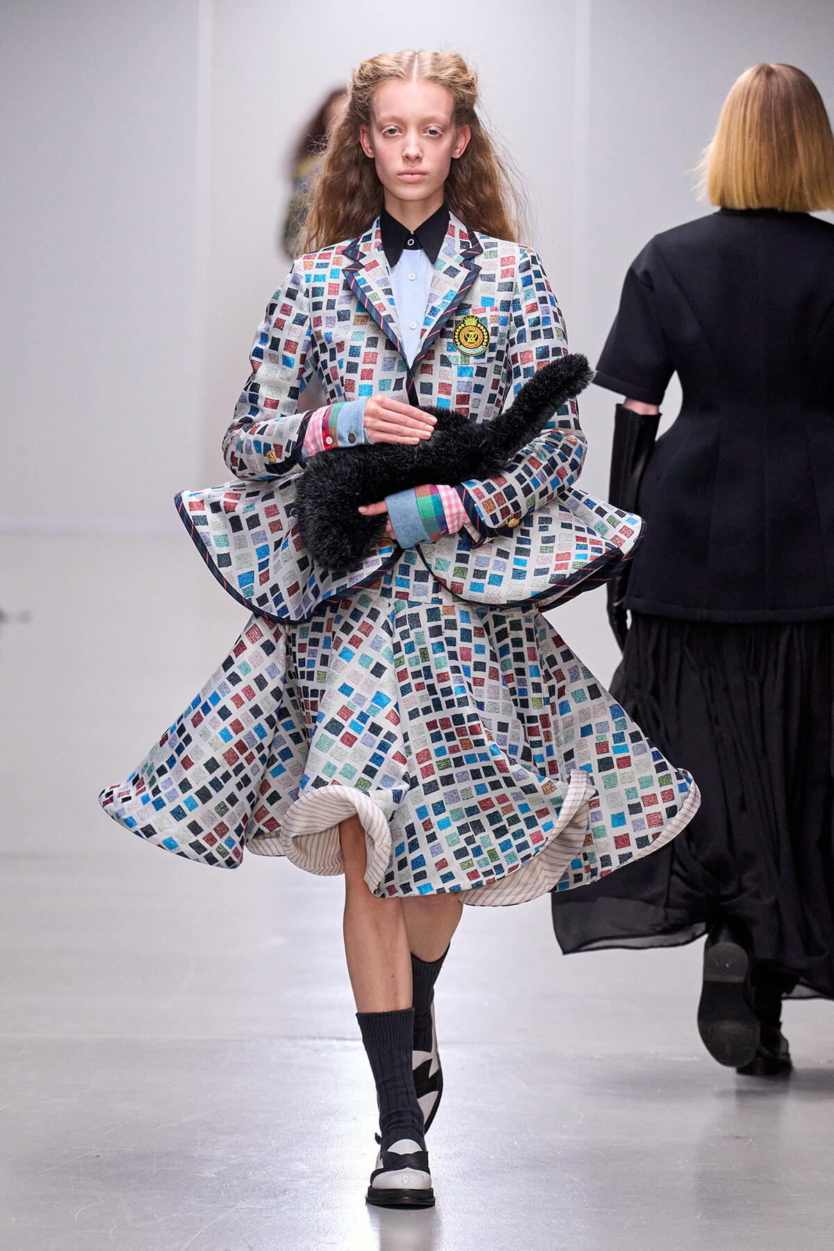 Model on a runway wearing a multicolored mosaic-pattern suit and dress, holding a black fluffy scarf with a badge on the chest.