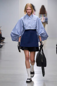 Model walks a fashion runway in a light blue striped blouse with oversized puff sleeves, paired with a navy short skirt, white socks, and black-and-white shoes while carrying a black bag and wearing metallic gloves.