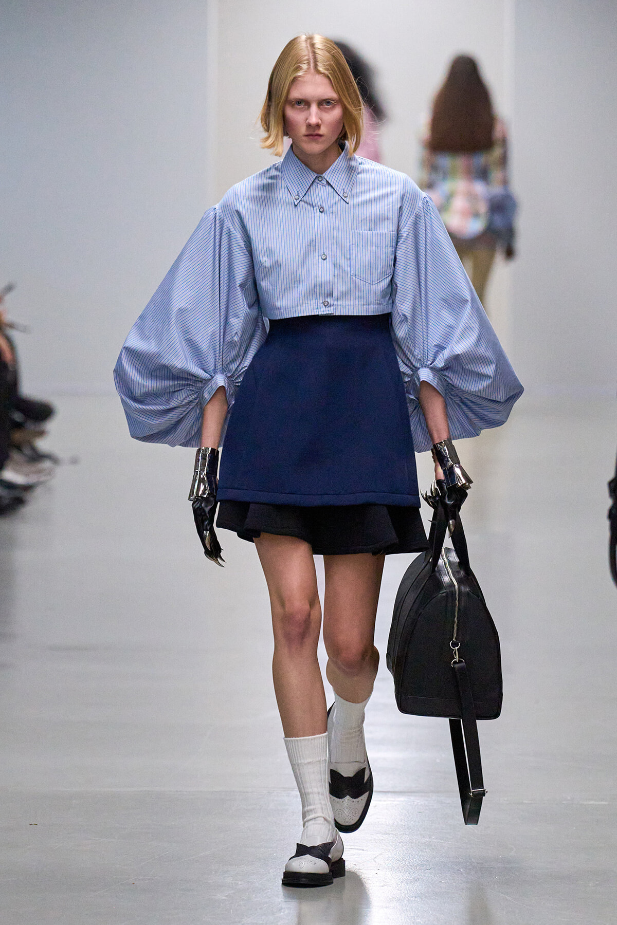 Model walks a fashion runway in a light blue striped blouse with oversized puff sleeves, paired with a navy short skirt, white socks, and black-and-white shoes while carrying a black bag and wearing metallic gloves.