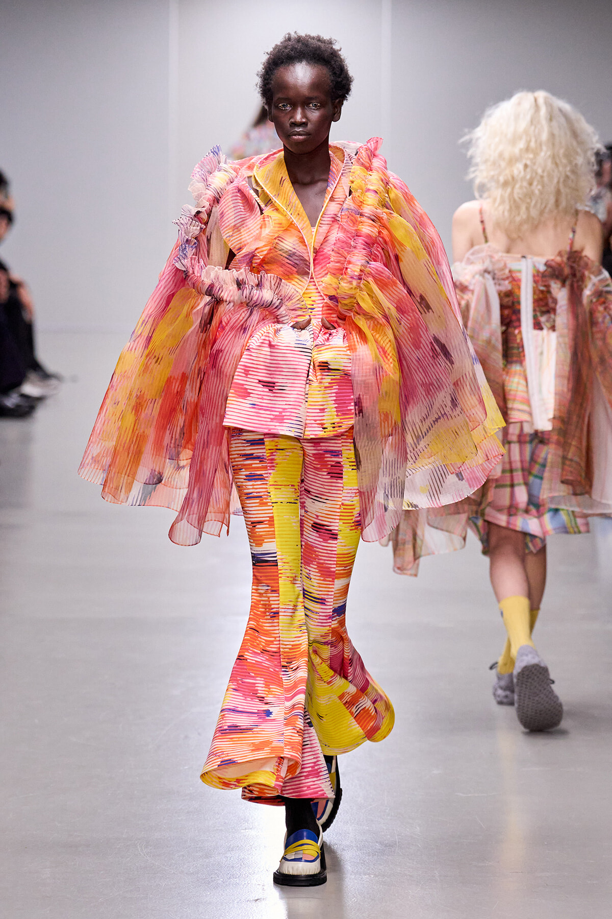 Model walking the runway in a bright, abstract pink, yellow, and orange ensemble with a flowing cape and matching patterned pants.
