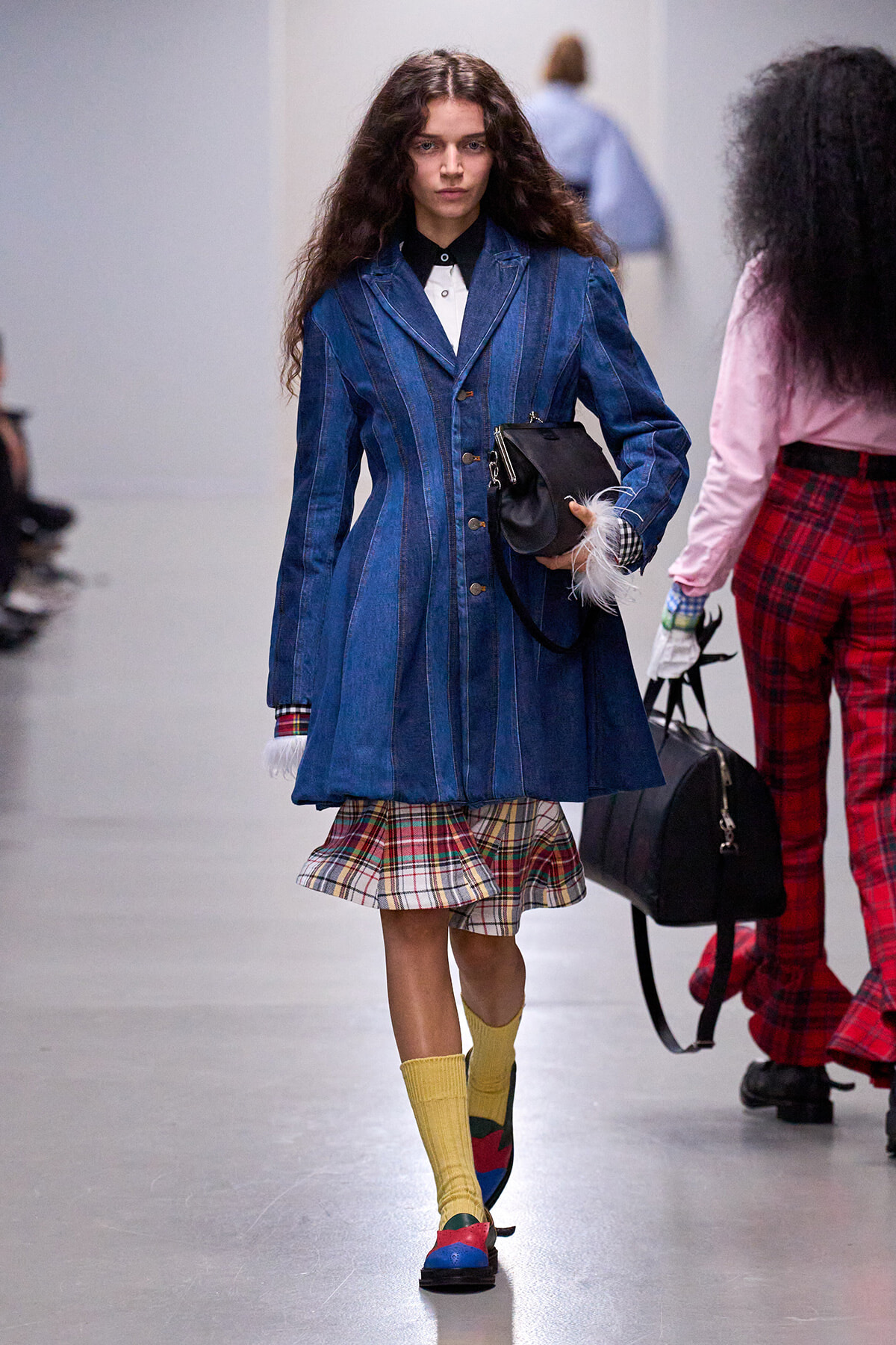 Model walking a runway in a blue striped denim coat over a plaid skirt, bright yellow knee-length socks, and multicolored loafers, carrying a black handbag.