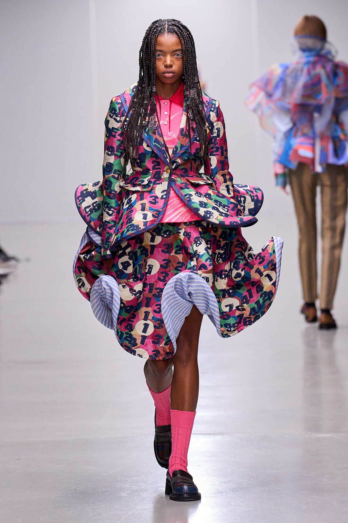Model walking the runway in a bold, multicolored abstract print suit with pink shirt and bright pink socks, black loafers visible.