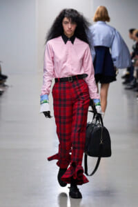 Model walks a runway in a pink button-up shirt with a black collar, red plaid pants, and black boots, carrying a black duffel bag.