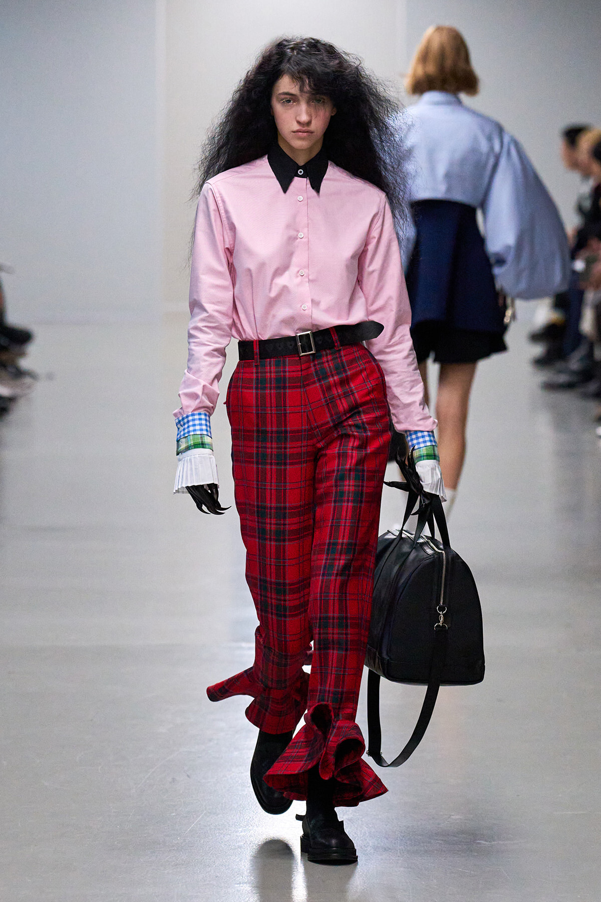 Model walks a runway in a pink button-up shirt with a black collar, red plaid pants, and black boots, carrying a black duffel bag.