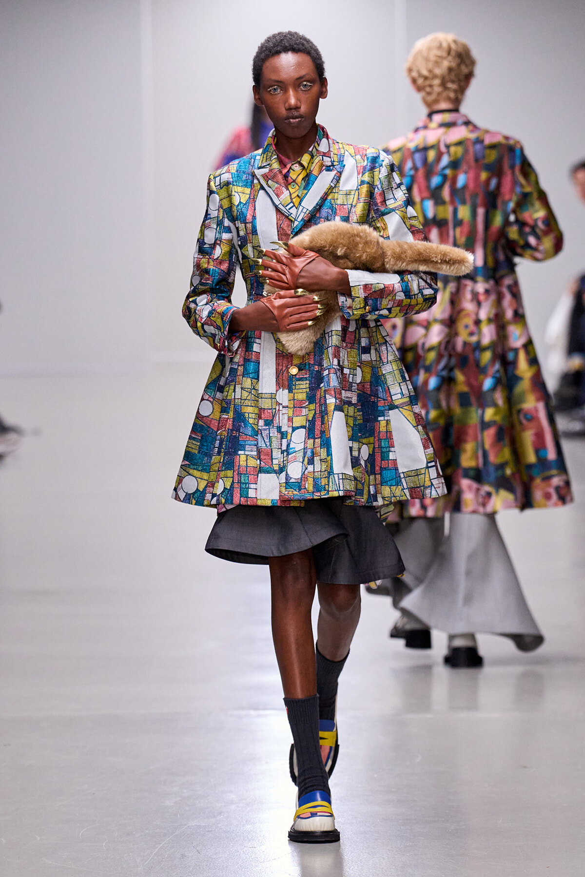 Model on a runway wearing a vibrant geometric-print coat, brown gloves, and a fur stole tucked under her arm.