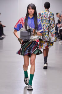 Model walking the runway in a patchwork blue top, multicolored striped pleated skirt, gray bag, green socks, and black‑white shoes with gold bracelets and gloves.