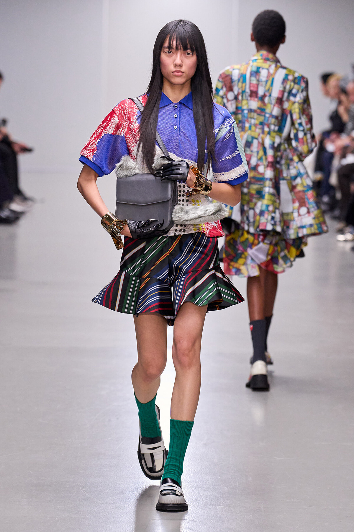 Model walking the runway in a patchwork blue top, multicolored striped pleated skirt, gray bag, green socks, and black‑white shoes with gold bracelets and gloves.