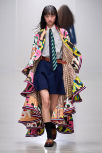 Model walking a runway in a vibrant, abstract-print cape layered over a white shirt, striped tie, and navy shorts.