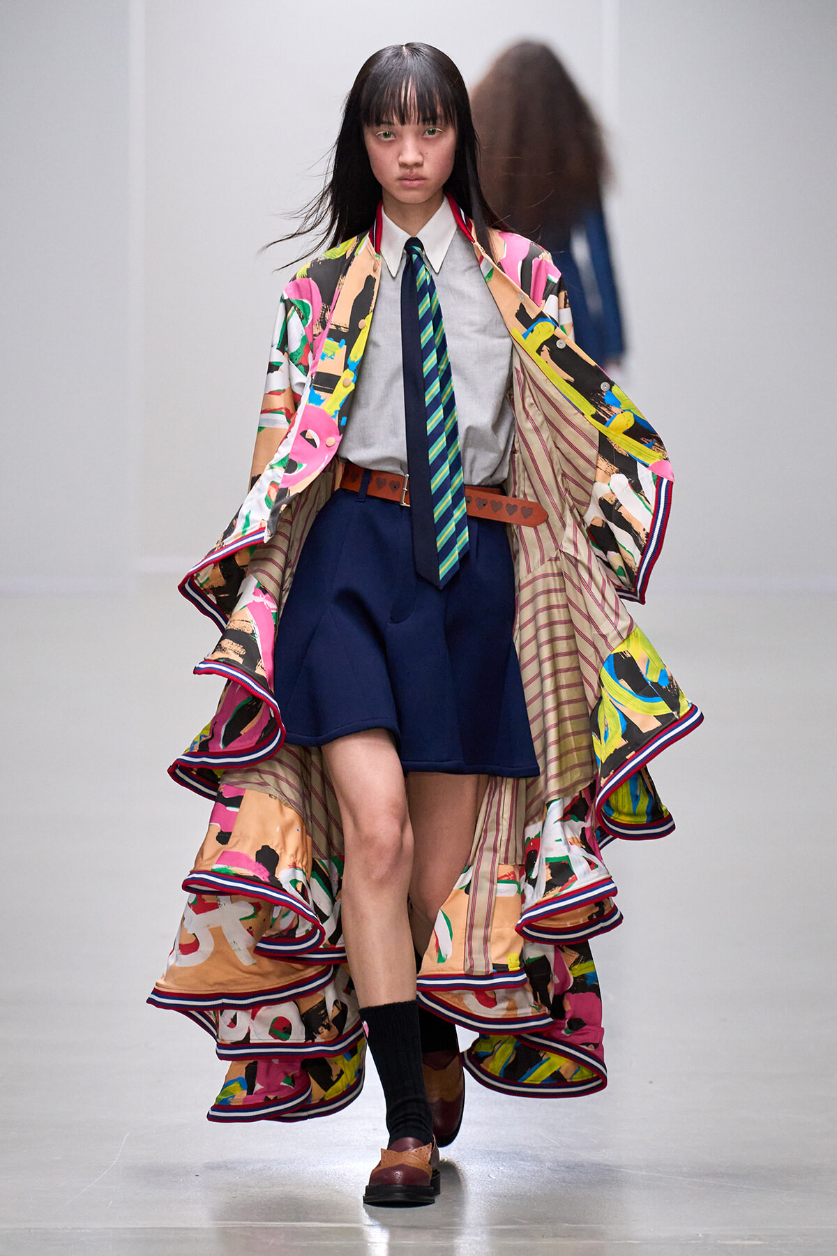 Model walking a runway in a vibrant, abstract-print cape layered over a white shirt, striped tie, and navy shorts.