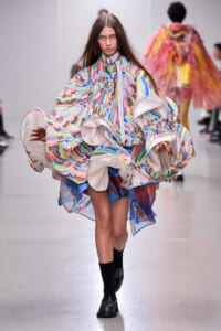 Model walking a fashion show in a multicolor, oversized coat with large circular ruffled sleeves and a striped interior lining.