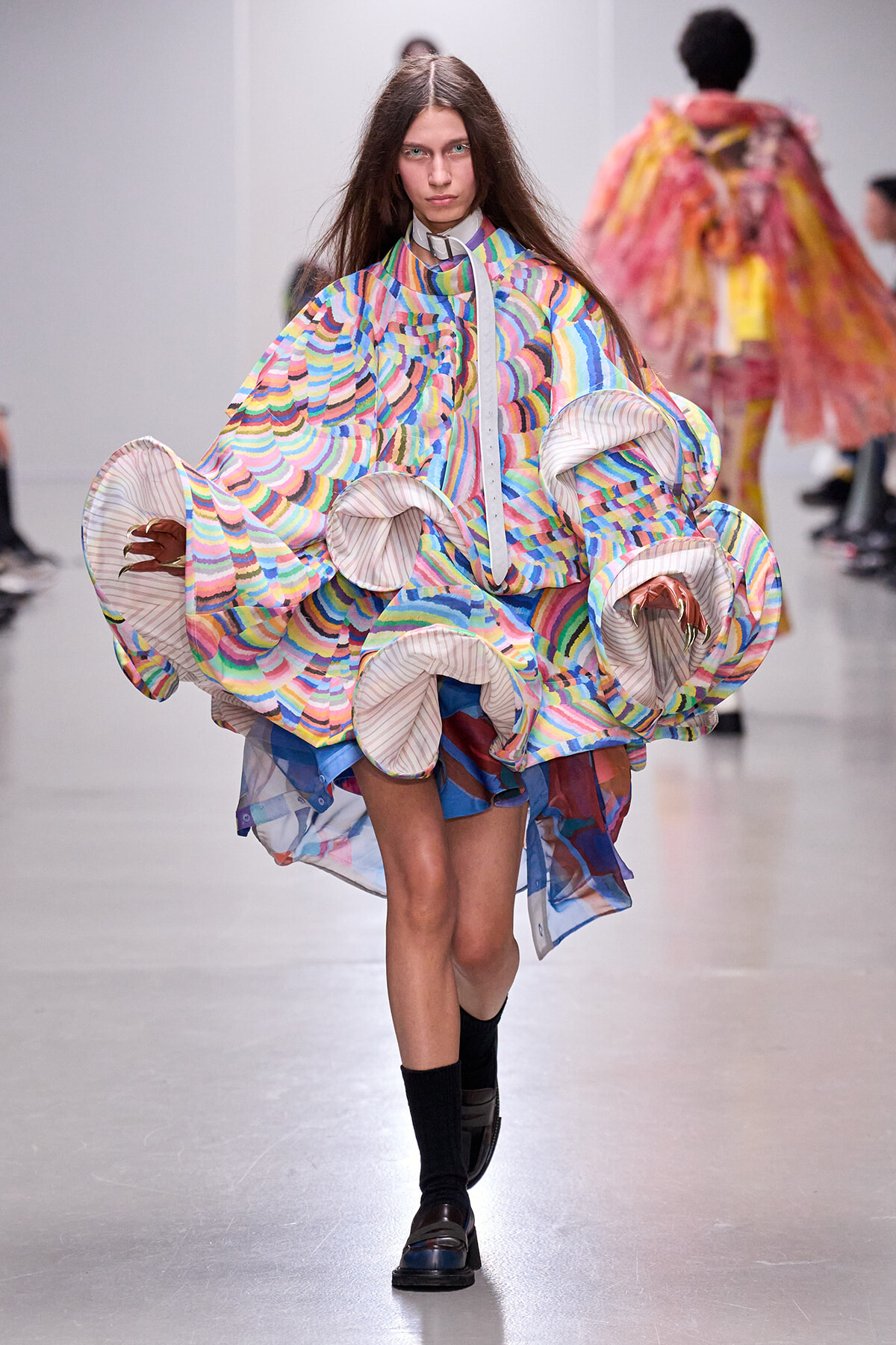 Model walking a fashion show in a multicolor, oversized coat with large circular ruffled sleeves and a striped interior lining.